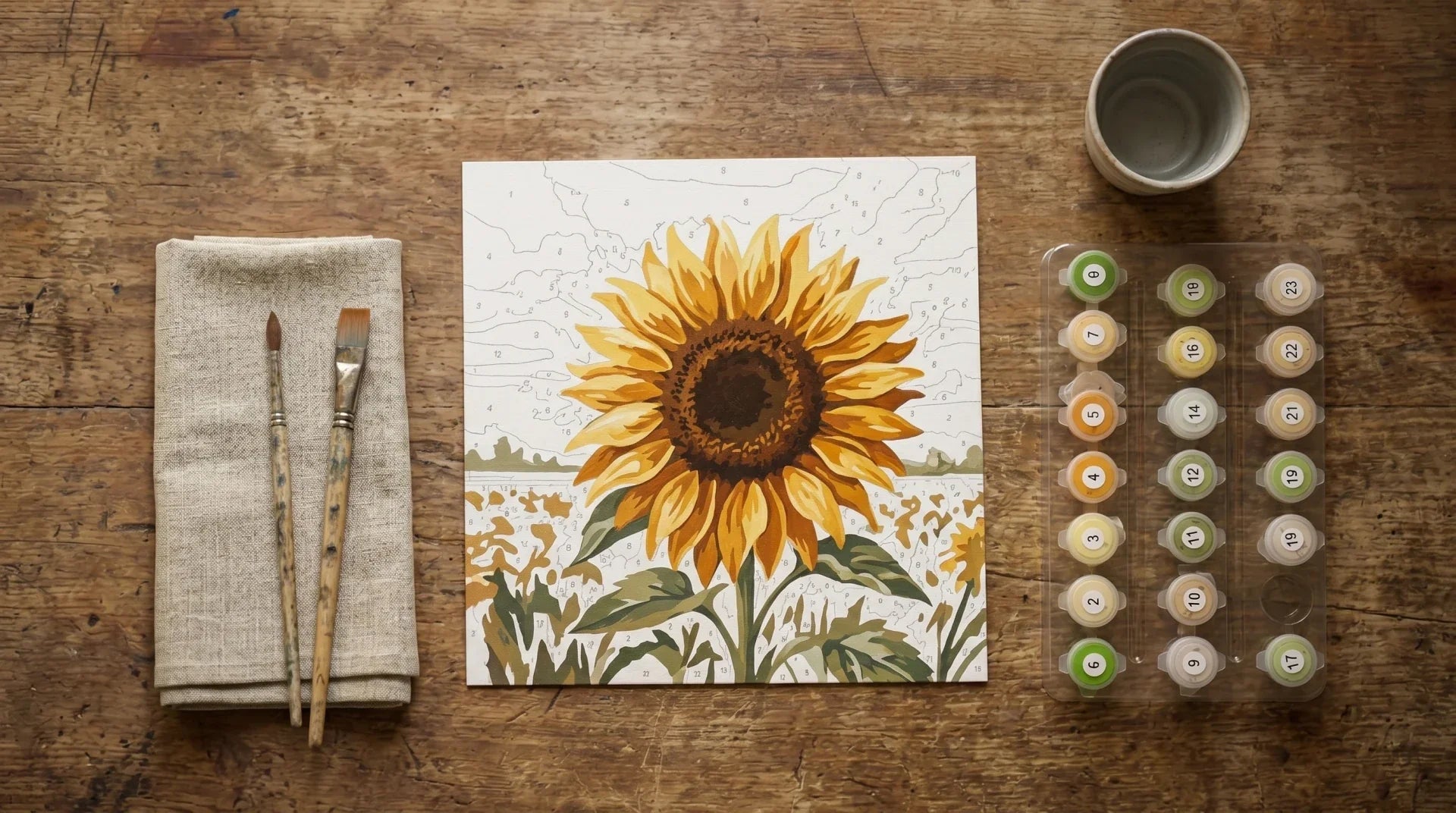 Paint by numbers canvas of a sunflower partly painted on a wooden table beside brushes and a numbered paint pot tray, in soft window light