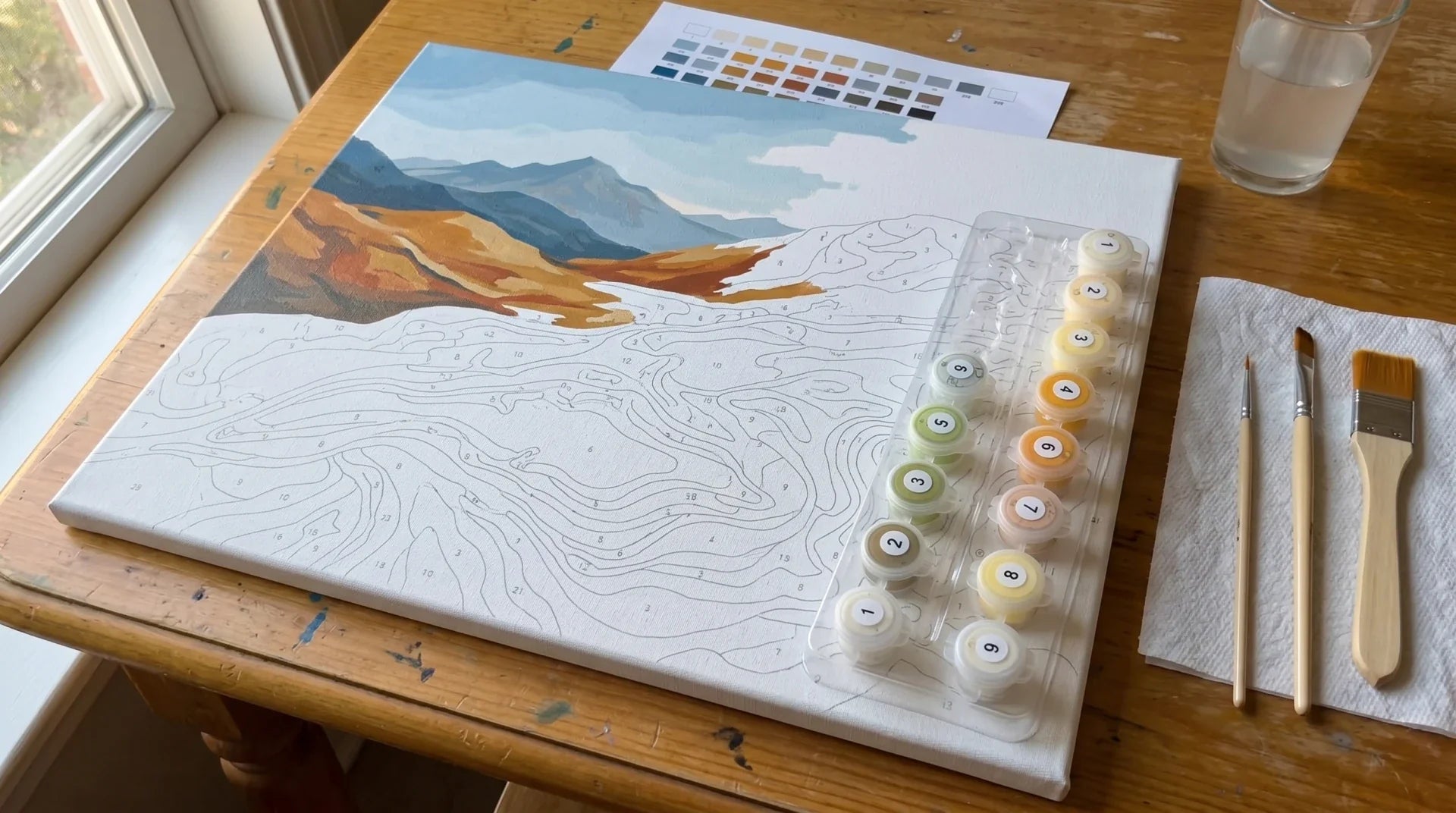 Top-down view of a paint by numbers workspace: a 16x20 canvas with a partially-painted mountain landscape, a clear plastic tray of connected numbered paint pots, three wooden-handled brushes, and a color reference sheet on a wooden table.