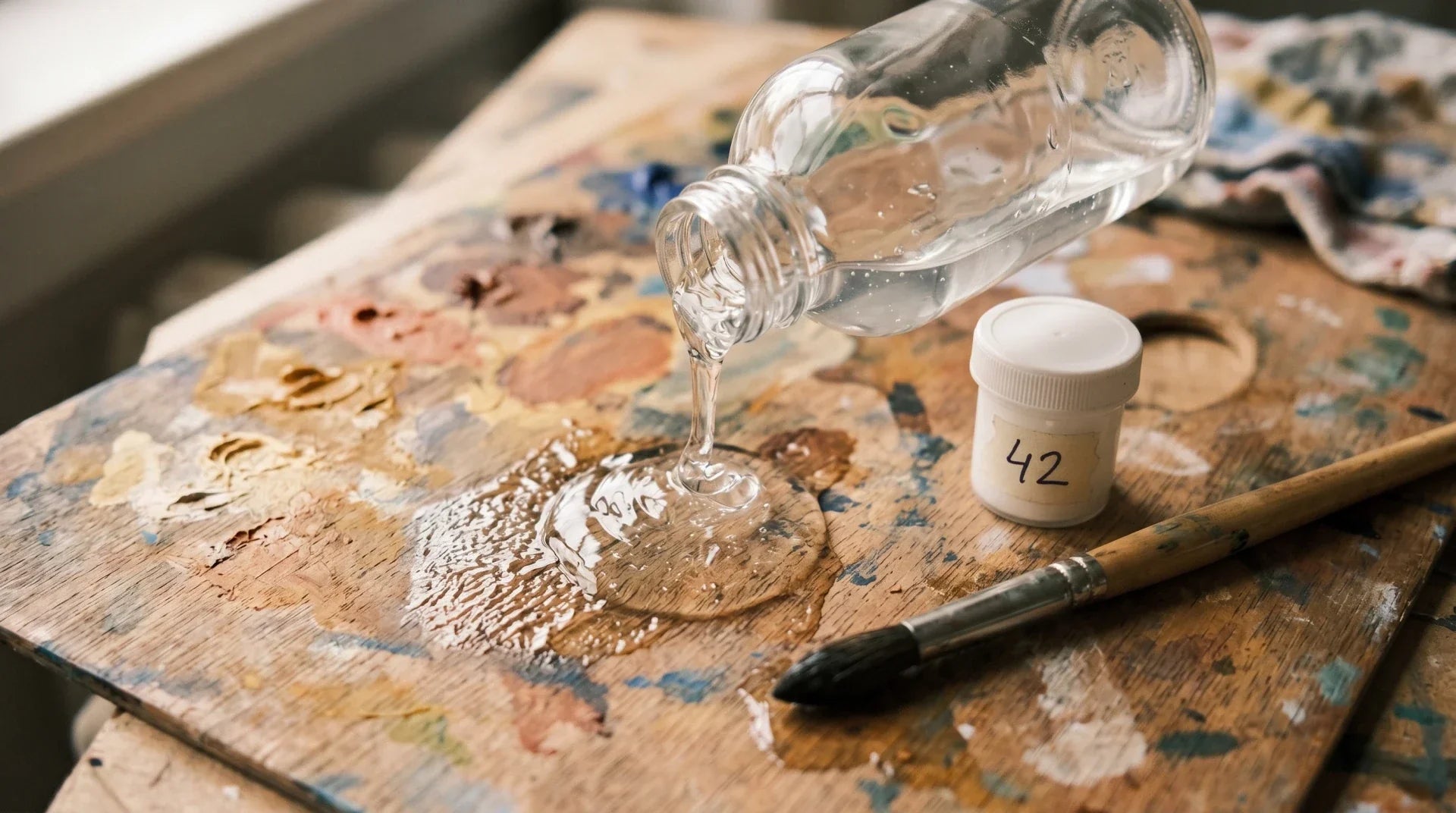 Unlabeled clear glass bottle pouring transparent acrylic gel medium onto a wooden artist palette, with a numbered paint pot 42 and round brush nearby.