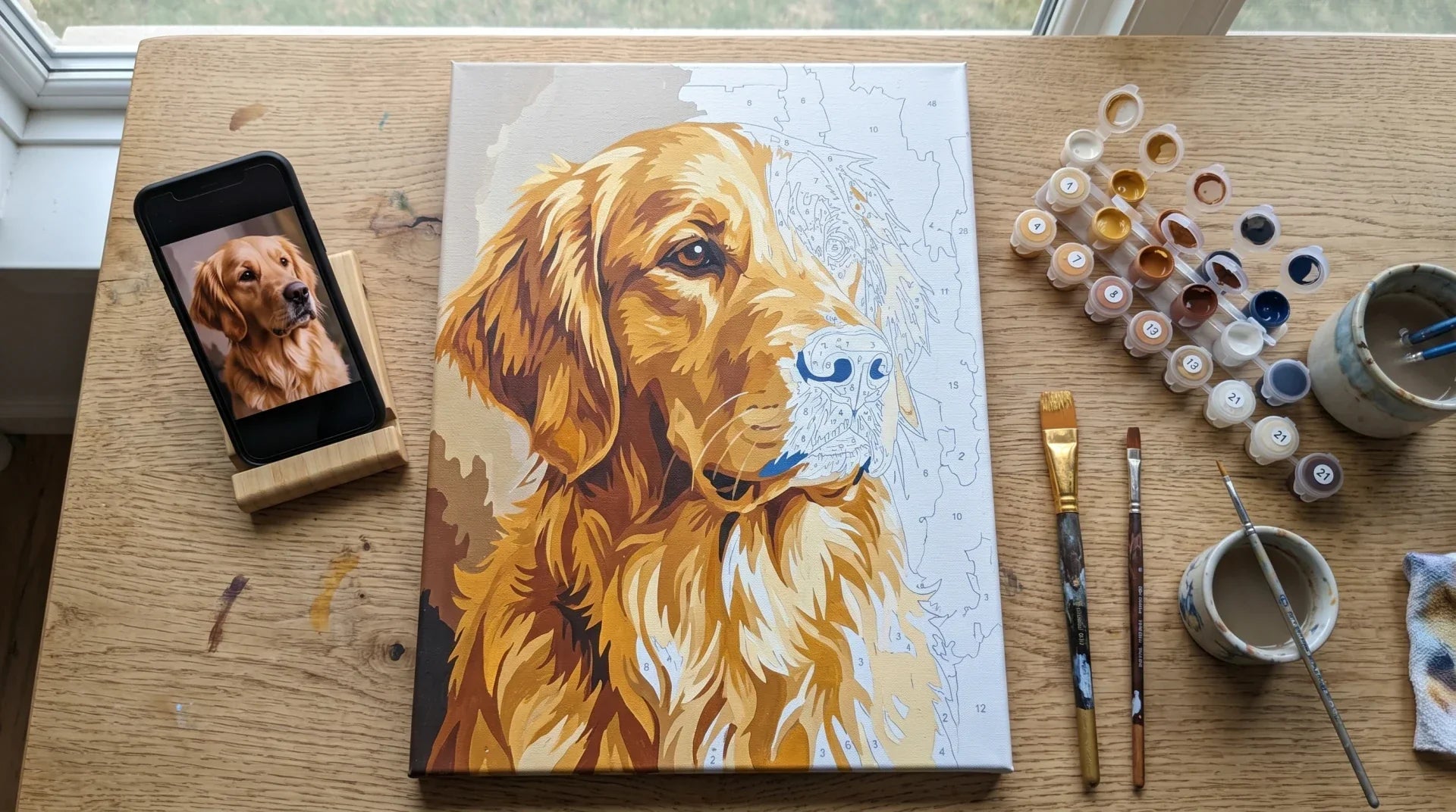 Overhead front view of a partly painted custom paint-by-numbers golden retriever on a gallery-wrapped stretched canvas with a phone showing the reference photo, numbered pots and brushes