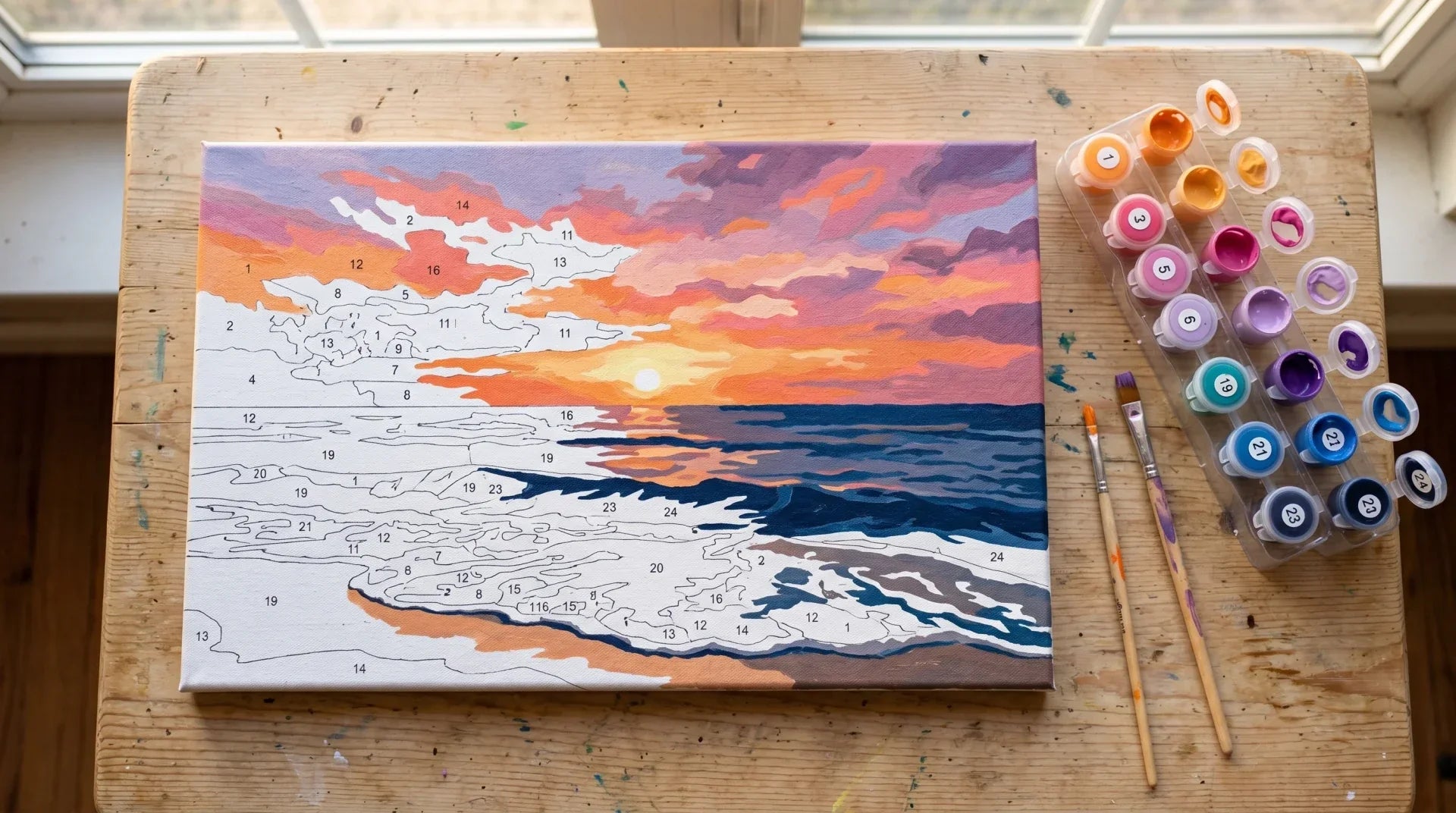 Overhead view of a partially painted paint-by-numbers canvas of a beach sunset with orange and pink sky and connected numbered pots beside it