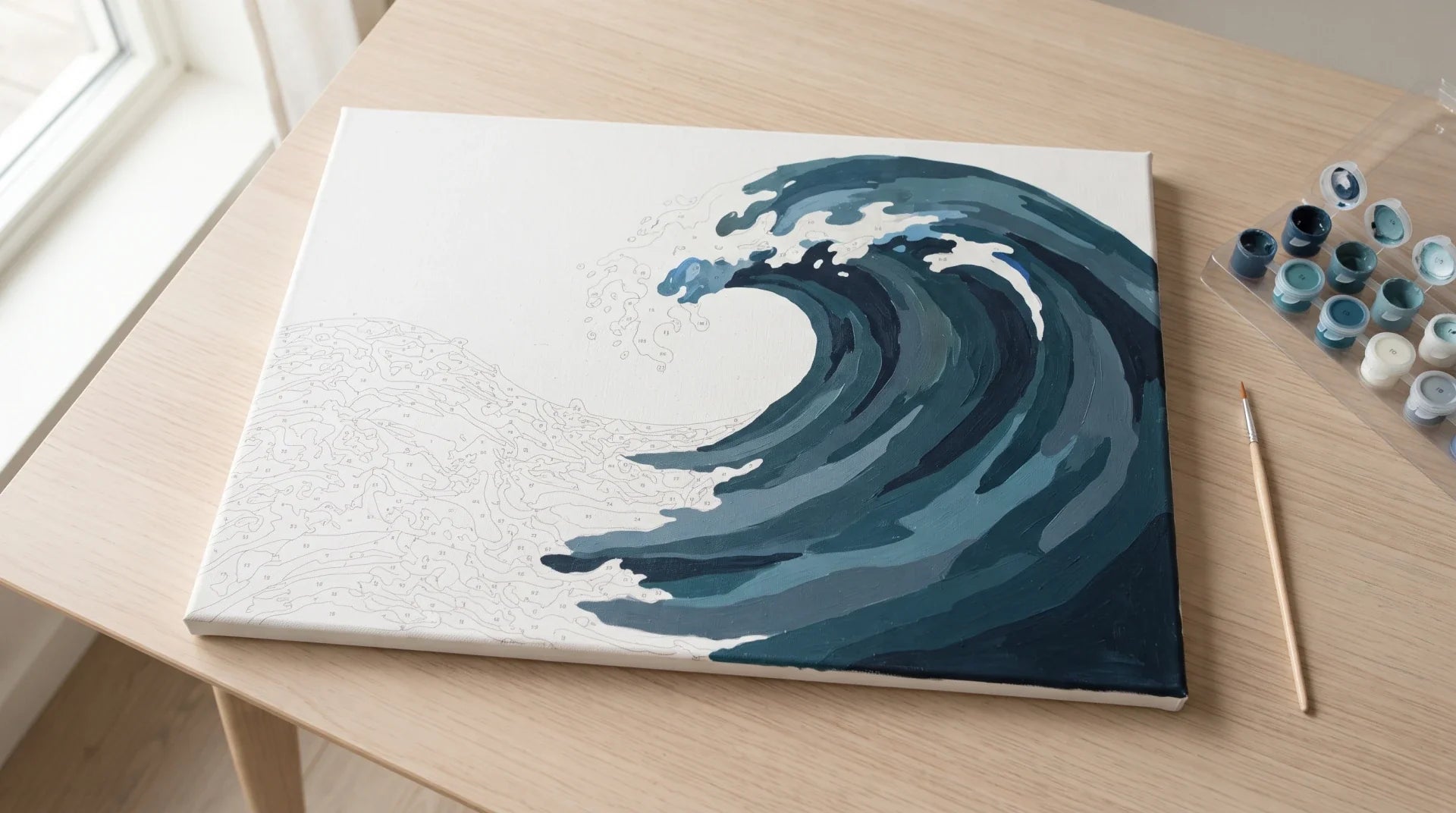 A part-finished paint by numbers canvas of a curling ocean wave on a light oak table, the unpainted foam still showing its printed outlines, with numbered paint pots and a brush alongside