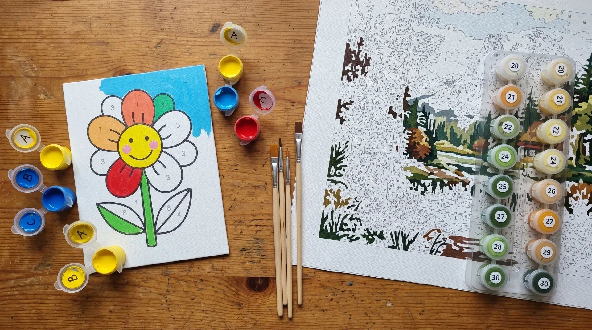 Top-down comparison of a small kid paint-by-numbers kit on the left and a larger adult paint-by-numbers landscape kit on the right, with brushes between them on a wooden table