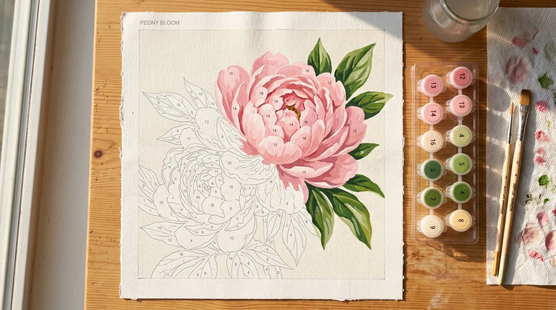 Overhead view of a partially painted paint-by-numbers canvas of a pink peony bloom with connected numbered paint pots and brushes on a warm wood table