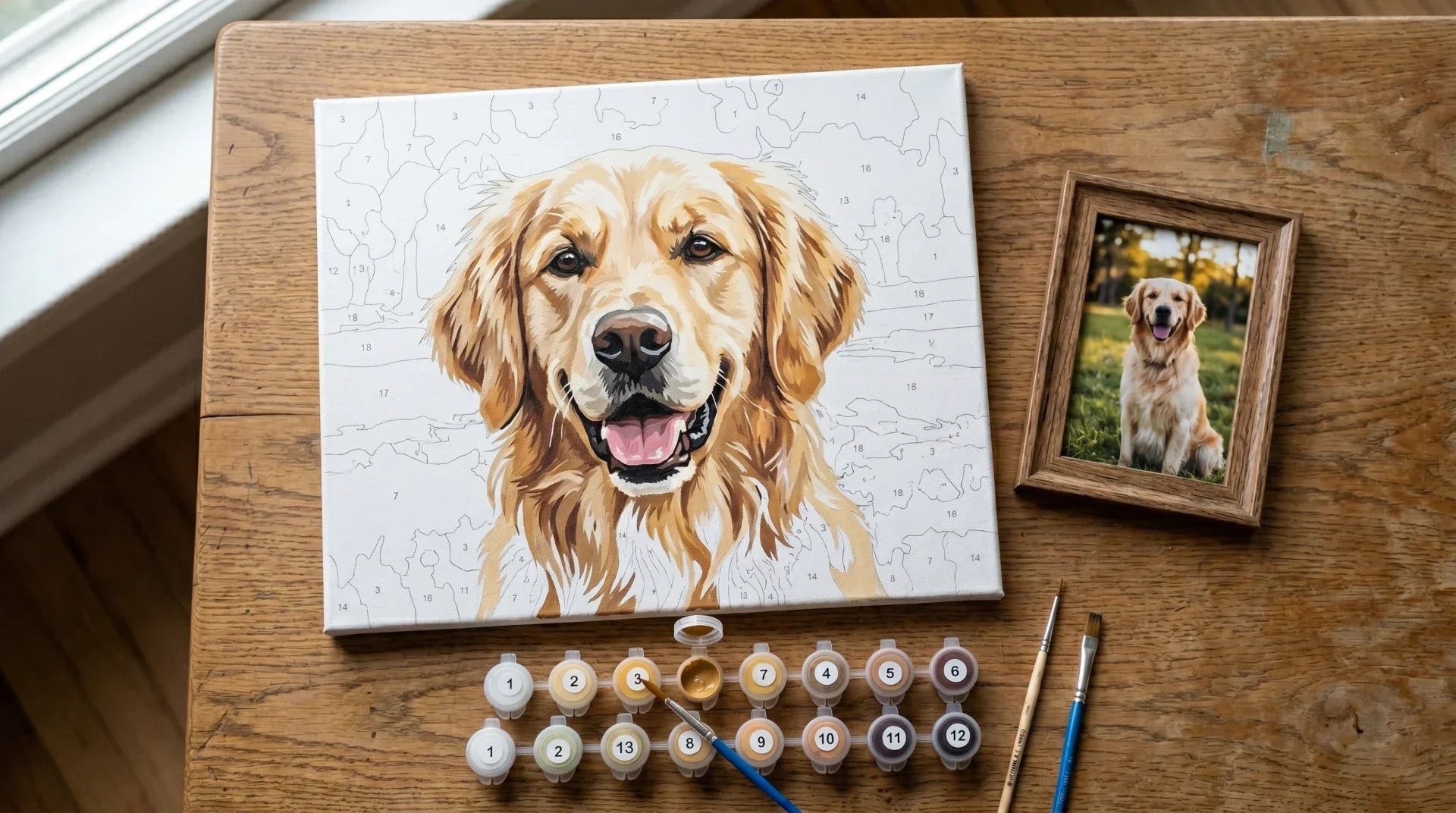 Partially painted paint-by-numbers canvas of a golden retriever portrait beside a small framed reference photograph of the same dog on a wooden table with paint pots and brushes
