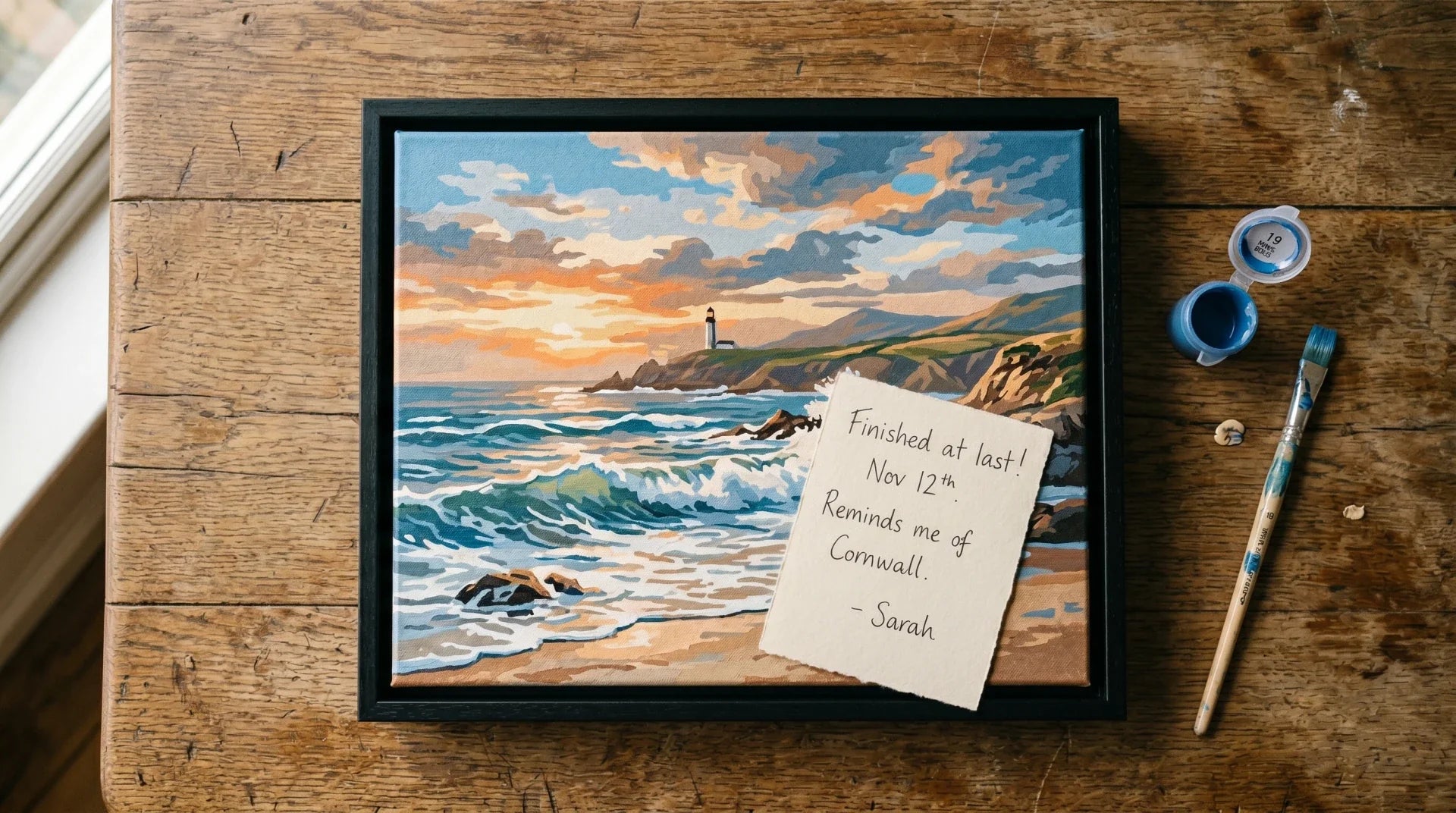 Top-down view of a finished paint by numbers seascape canvas in a black floating frame on a worn wooden table, with a small handwritten card on top, plus a numbered paint pot and brush beside it in soft warm window light.