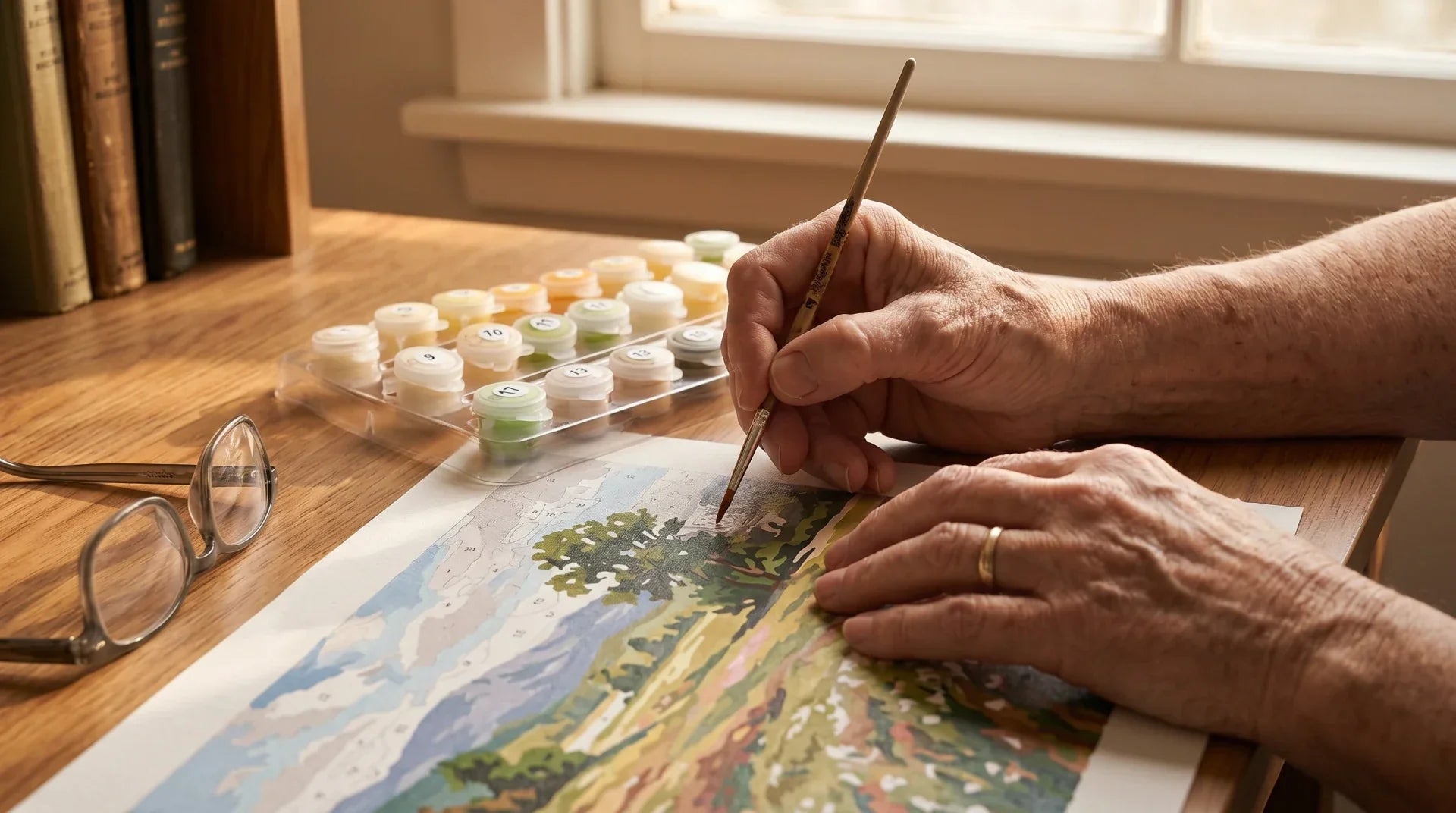 Close-up of an older adult's hands holding a fine brush, painting a paint-by-numbers landscape on a wooden table with reading glasses and a numbered paint pot tray in warm afternoon light