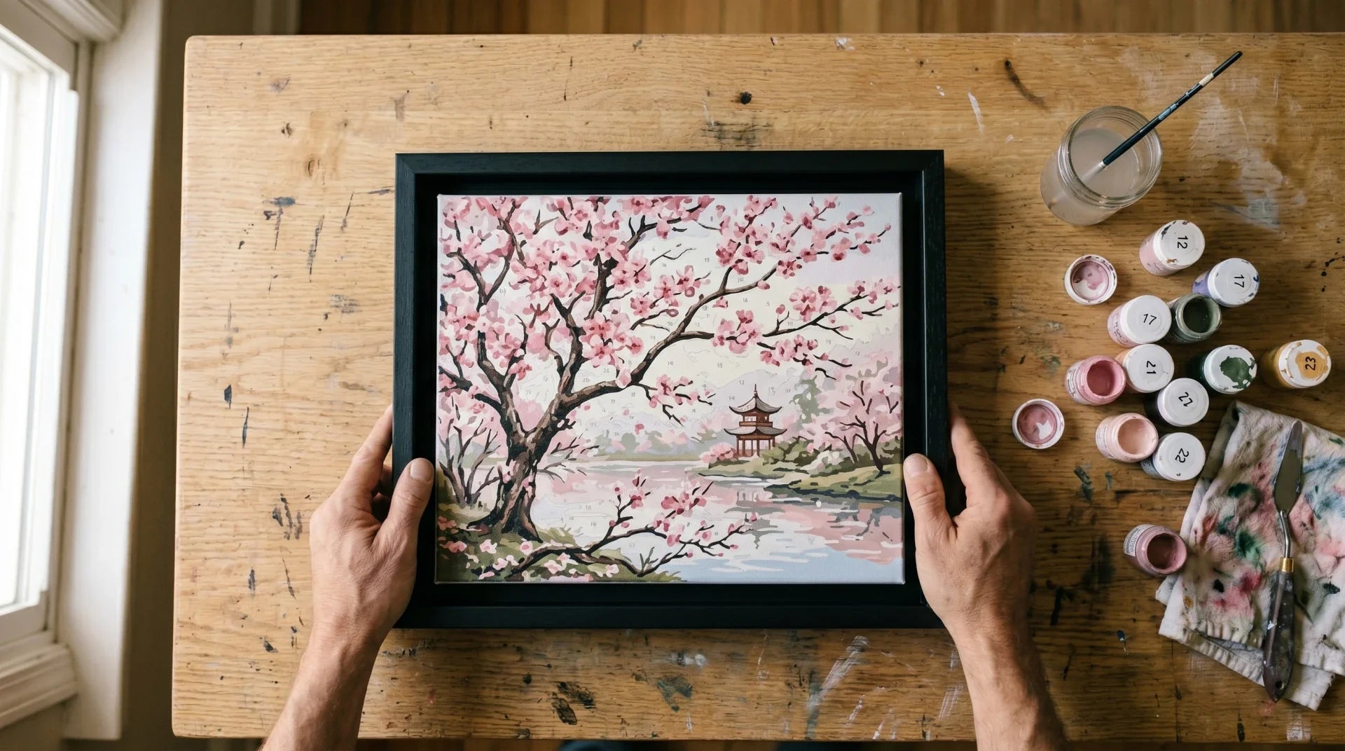 Top-down view of a finished paint-by-numbers cherry blossom canvas being placed into a matte black floating frame on a worn light oak workshop bench, with soft natural side window light and a few numbered paint pots resting nearby.
