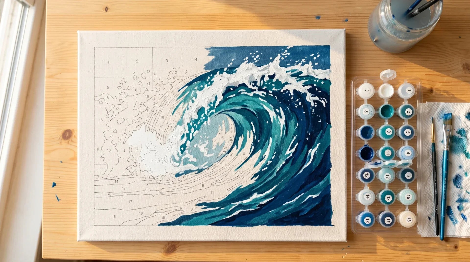 Overhead view of a partially painted paint-by-numbers canvas of a crashing ocean wave with connected numbered pots and brushes on a wood table
