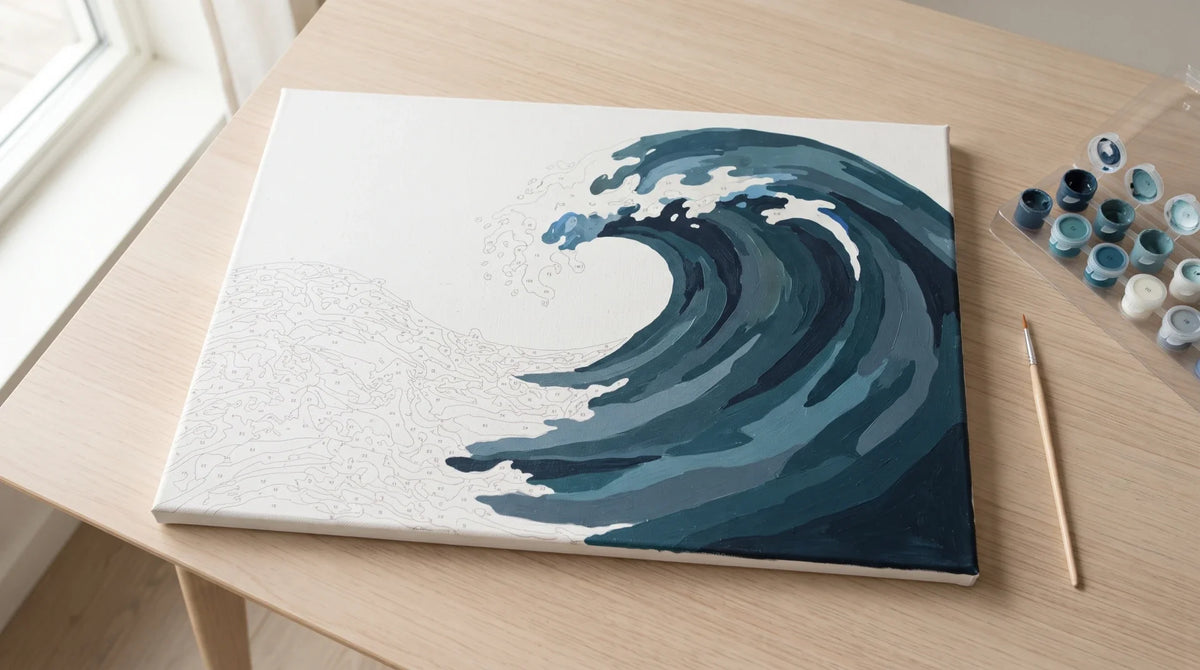 A part-finished paint by numbers canvas of a curling ocean wave on a light oak table, the unpainted foam still showing its printed outlines, with numbered paint pots and a brush alongside