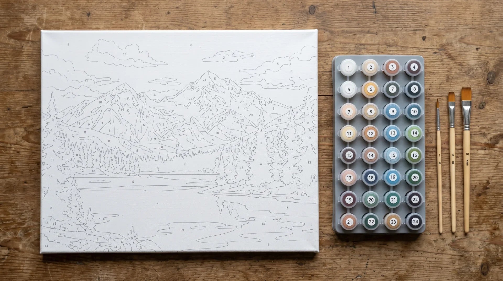 A paint-by-numbers kit stored on a table: a gallery-wrapped stretched canvas printed with a mountain-lake landscape, a tray of numbered paint pots with lids shut, and brushes