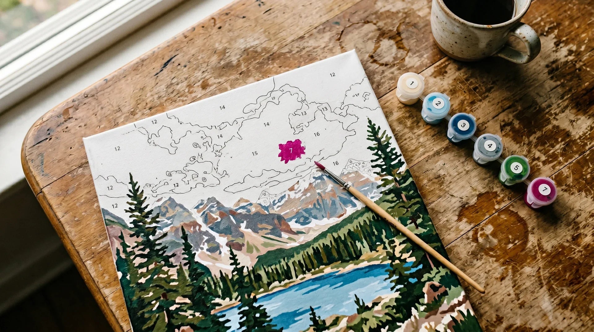 Half-finished paint by numbers mountain landscape canvas with a small magenta mistake in the cloud area, alongside numbered paint pots and a brush on a wooden table.