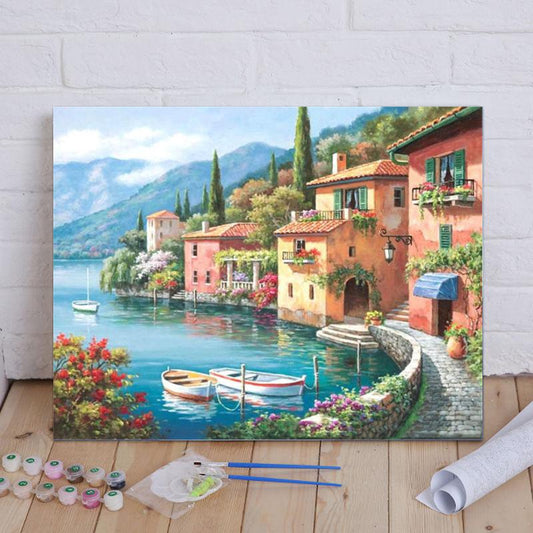 Painting of a scenic Italian village by a lake with boats and flowers on a wooden floor.