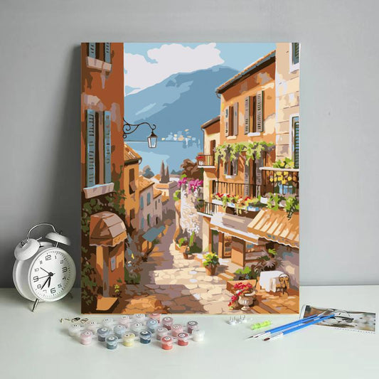 Painting of a scenic street with mountains in the background, displayed on a gray surface with art supplies.