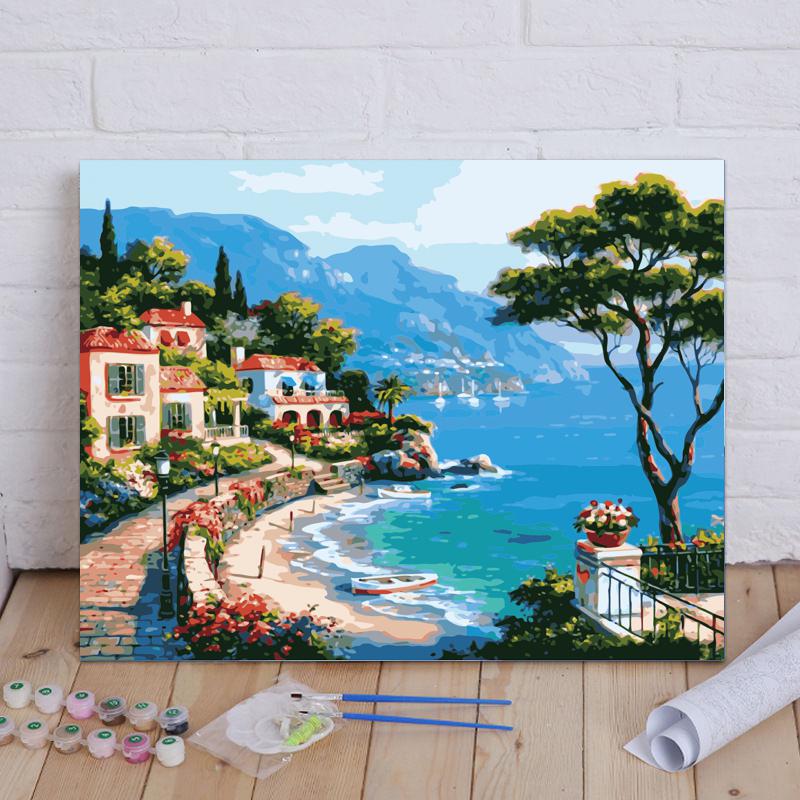 Painting of a coastal scene with houses, trees, and mountains on a wooden floor.